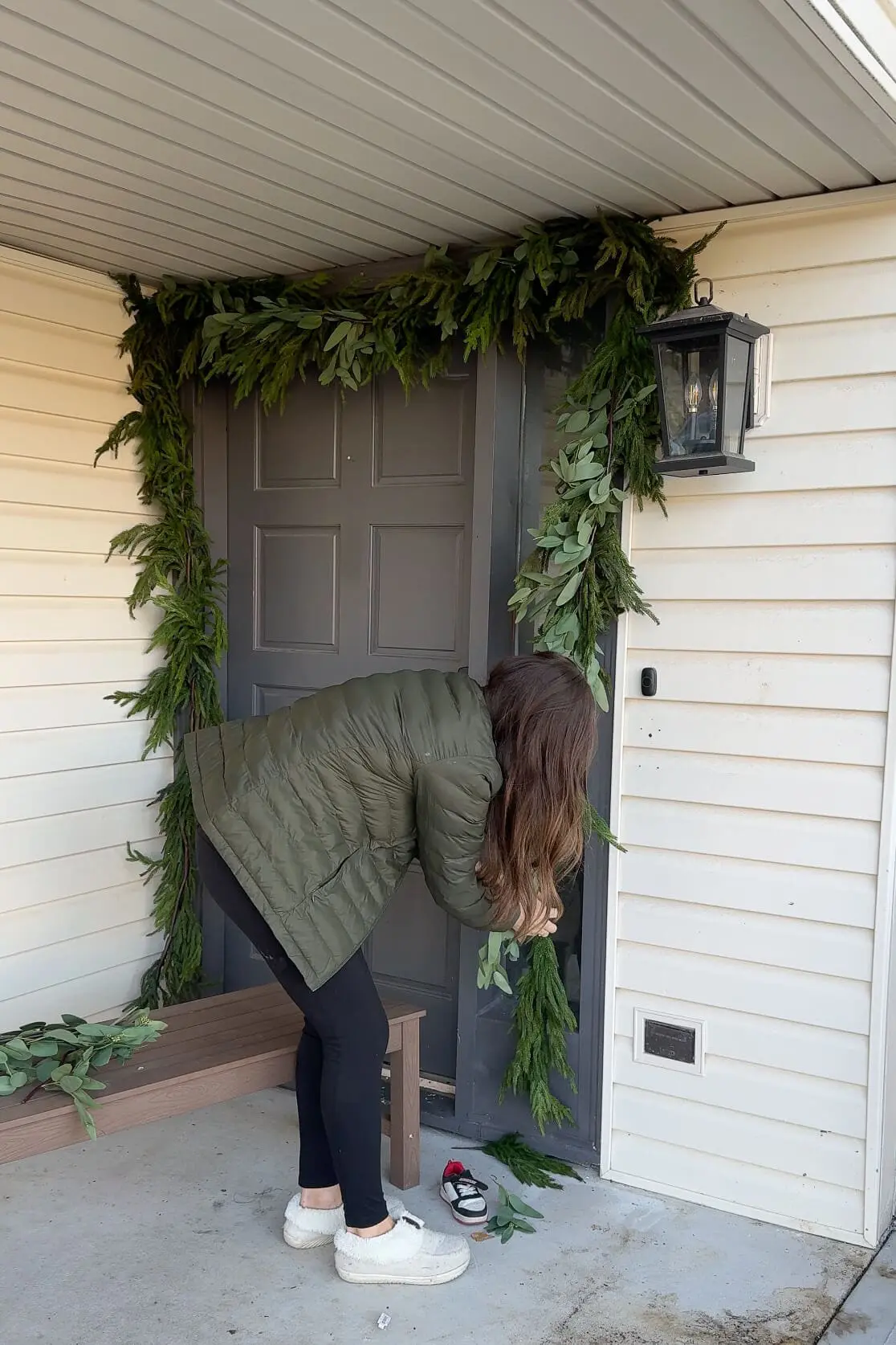 A woman in a green puffer jacket and black leggings carefully arranging fresh greenery garland around a gray front door, with a bench nearby and white siding framing the entrance.